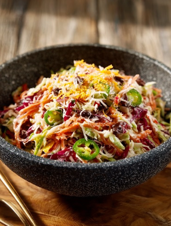 Bowl of colorful cowboy coleslaw featuring shredded cabbage, carrots, sliced jalapeños, and dried cranberries, garnished with grated cheese