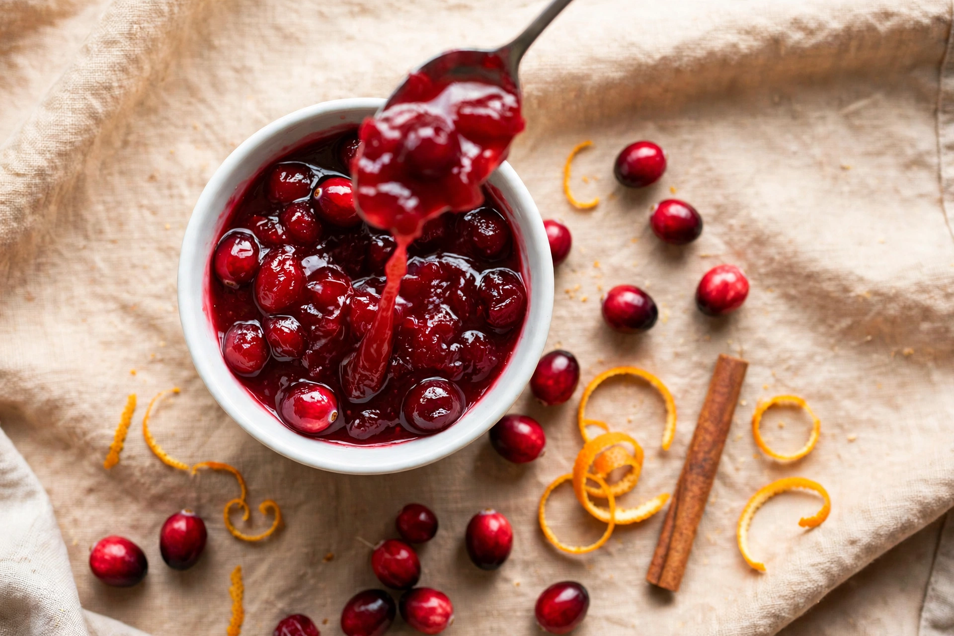 Make-ahead cranberry orange sauce in a white bowl, a spoon lifting a glossy ribbon of sauce, whole berries, orange zest curls and a cinnamon stick beside the bowl